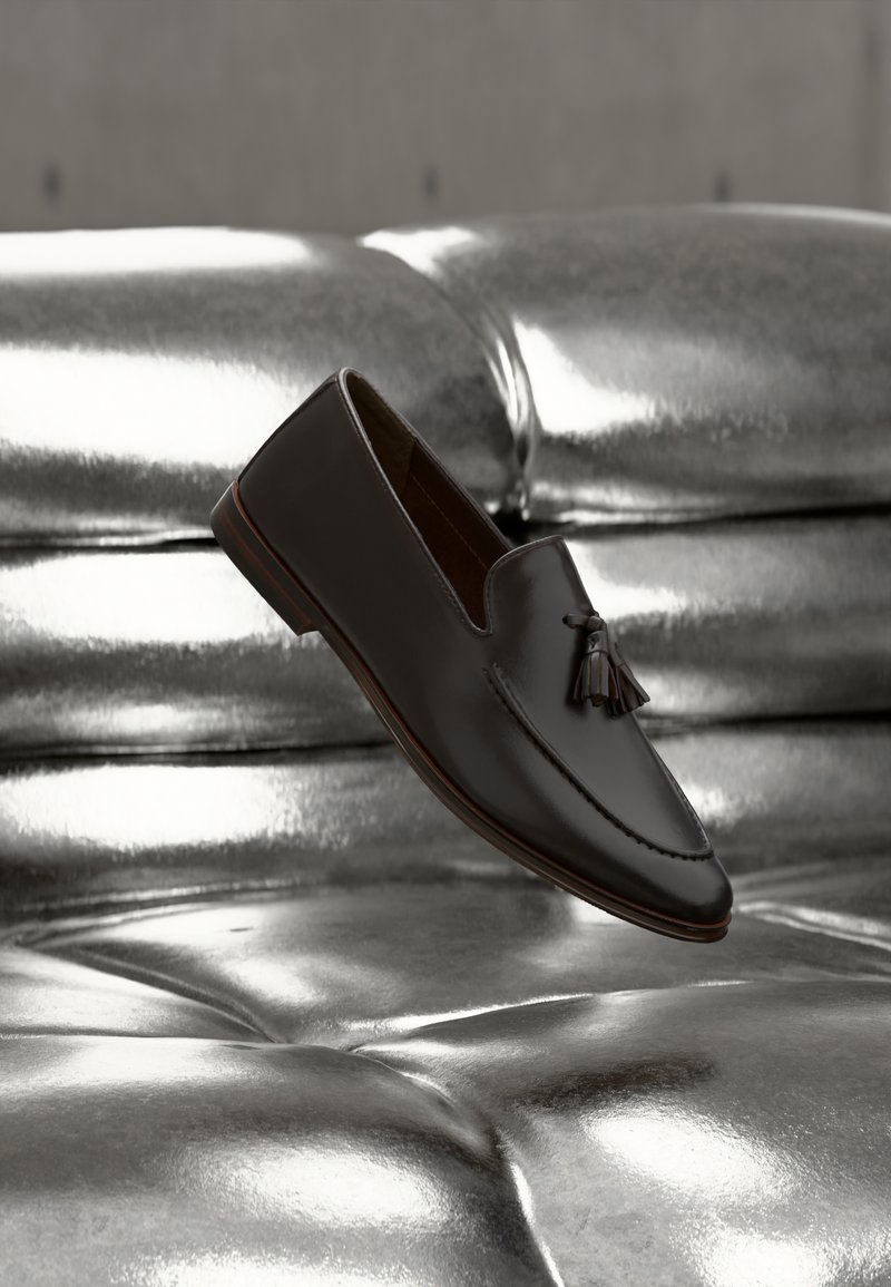 Mocassin à pampilles en cuir brun foncé flottant au-dessus d'un canapé argenté brillant et capitonné avec une surface réfléchissante.
