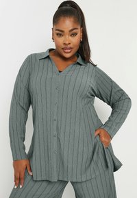 Camicia grigia a maniche lunghe con bottone e scollatura a V, caratterizzata da un motivo a coste testurizzato e una vestibilità rilassata. Il materiale sembra morbido e leggero.