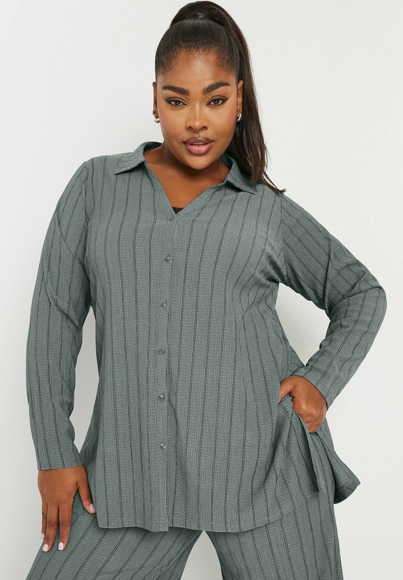 Camicia grigia a maniche lunghe con bottone e scollatura a V, caratterizzata da un motivo a coste testurizzato e una vestibilità rilassata. Il materiale sembra morbido e leggero.