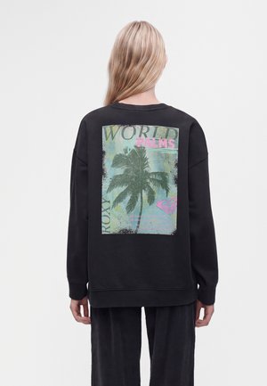 Personne aux longs cheveux blonds portant un sweat-shirt noir avec un grand motif de palmier et les mots "WORLD PALMS" au dos.