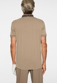 Mężczyzna zwrócony tyłem, ubran w beżową koszulkę polo z krótkim rękawem, z czarnym, wzorzystym wykończeniem kołnierza, beżowe spodnie oraz białą rękawiczkę na lewej ręce.