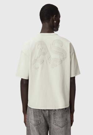 Person trägt ein locker sitzendes, cremefarbenes Kurzarmhemd mit strukturierten "AS"-Buchstaben auf dem Rücken und ausgewaschene graue Jeans, steht vor einem schlichten Hintergrund.