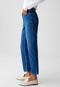 Personne portant un jean droit taille haute bleu, un haut blanc et des mocassins crème, les mains dans les poches devant, sur fond uni.