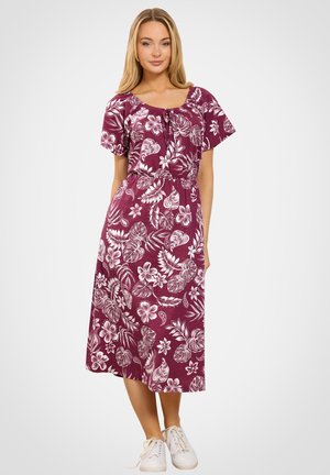 Kvinde stående iført en lilla midi-kjole med hvidt tropisk blomsterprint og hvide sneakers mod en ensfarvet lys baggrund.