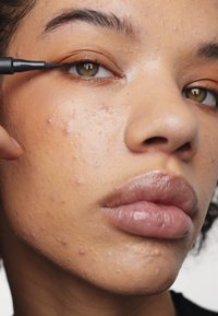 Eine Nahaufnahme eines Gesichts, das schwarzen flüssigen Eyeliner aufträgt, mit glatter, leicht fettiger Haut und kleinen Unreinheiten sowie vollen, glänzenden Lippen.