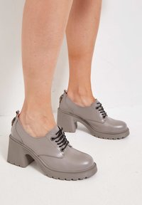 Zapatos de plataforma grises con un acabado brillante, cordones negros y un tacón grueso y texturizado. Presenta una punta redondeada y una pequeña lengüeta en el talón.