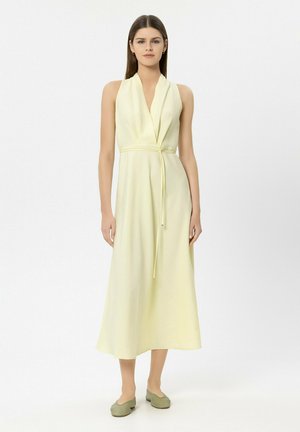 Frau mit glattem braunem Haar, die ein ärmelloses, blassgelbes Midikleid mit V-Ausschnitt und Bindegürtel trägt, dazu salbeigrüne Ballerinas.