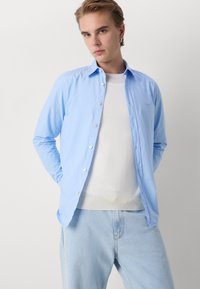 Camicia button-up azzurro chiaro con maniche lunghe, dotata di colletto a punta e bottoni bianchi, indossata sopra una maglietta bianca a girocollo.