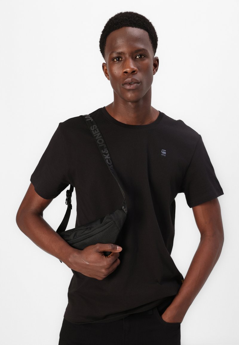 T-shirt noir en tissu lisse, avec un petit logo sur la poitrine. Le modèle tient un sac bandoulière noir avec une sangle à motif.