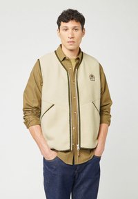Giacchetta in pile beige con rifiniture verdi, dotata di zip frontale e due grandi tasche. Indossata sopra una maglietta oliva e jeans scuri.