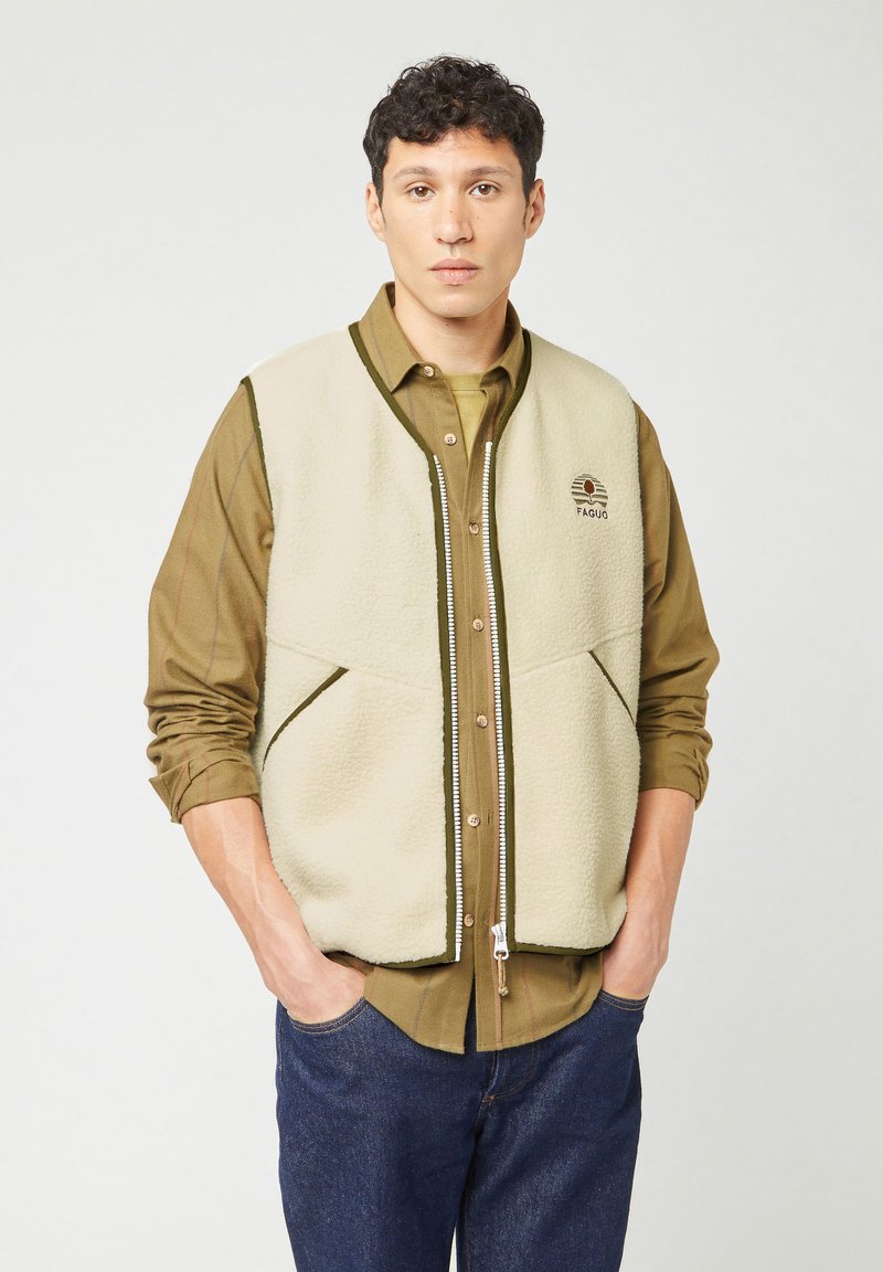 Giacchetta in pile beige con rifiniture verdi, dotata di zip frontale e due grandi tasche. Indossata sopra una maglietta oliva e jeans scuri.