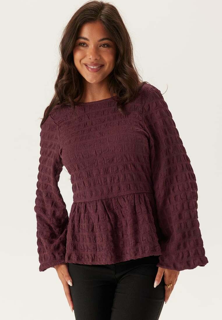 Bordeaux gekleurde, textuurvaste top met lange mouwen en een peplum taille, voorzien van een ronde halslijn en volants op de mouwen, gecombineerd met donkere broek.