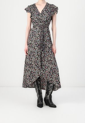 Femme portant une robe noire à fleurs avec des manches volantées et des bottes de cowboy noires brodées, debout contre un mur uni.