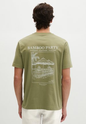 Man draagt een olijfgroen T-shirt met een witte schets van een tiki-bar, krukken, palmboom en tekst "Bamboo Party, Good Tunes, Good Drinks, Good Food."