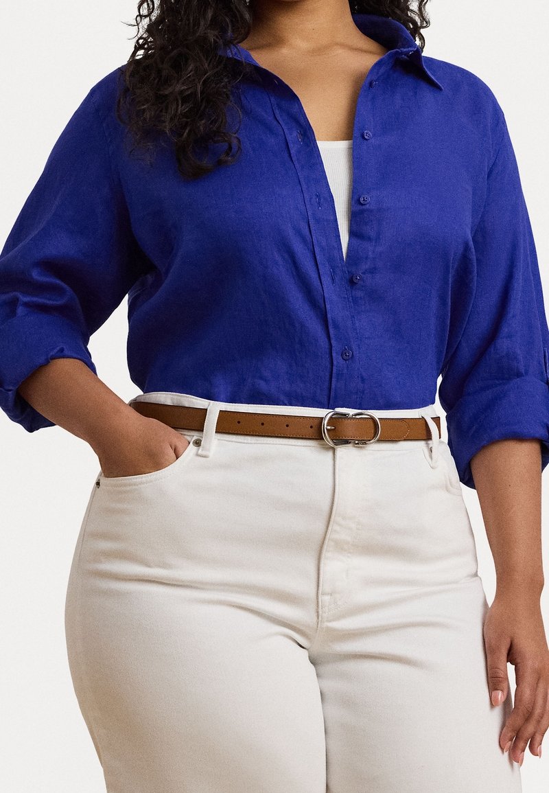 Femme portant une chemise bleue à boutons avec les manches retroussées par-dessus un haut blanc, rentrée dans un pantalon blanc taille haute avec une ceinture marron.