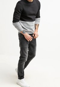 Hombre vestido con una camiseta de manga larga negra y gris, jeans negros y zapatillas blancas, ajustándose la cintura de los jeans, con tatuajes visibles en los antebrazos.