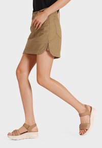 Gonna mini in denim khaki con orlo curvo e spacchi laterali, abbinata a sandali strappy marrone chiaro con soletta spessa e cinturini regolabili.