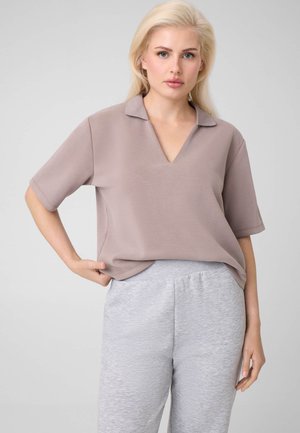 Femme blonde portant un haut ample mauve clair à manches courtes et un pantalon de survêtement gris clair, debout avec une main sur la hanche.