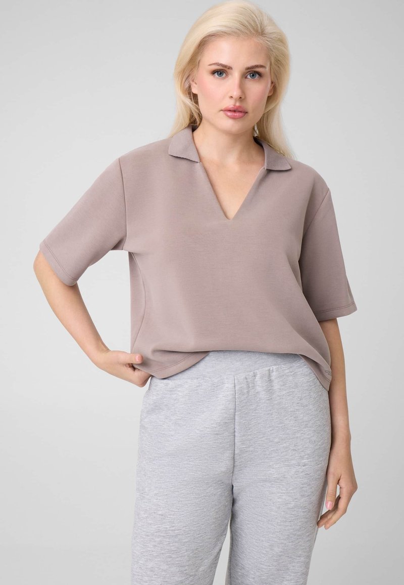 Femme blonde portant un haut ample mauve clair à manches courtes et un pantalon de survêtement gris clair, debout avec une main sur la hanche.