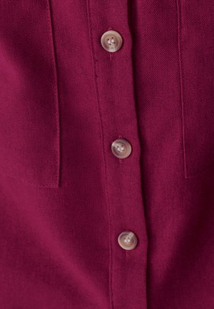 Fuksijaste bombažno srajco z odkrito zapenjanje s štirimi usklajenimi gumbi in dvema žepoma na prsih. Tkanina ima gladko teksturo.