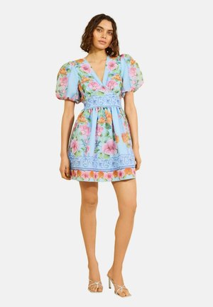 Frau trägt ein kurzes, hellblaues geblümtes Kleid mit Puffärmeln und V-Ausschnitt, kombiniert mit weißen Sandalen mit hohen Absätzen.