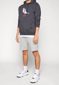 Sudadera gris con un gráfico de gaviota, bolsillo canguro y cordón. Combinada con pantalones cortos grises y zapatillas blancas con detalles en negro.