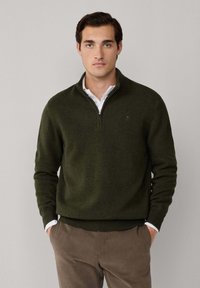 Suéter de lana verde con cuello tipo media cremallera, puños y bajo acanalados, superpuesto a una camisa blanca con cuello, combinado con pantalones marrones.