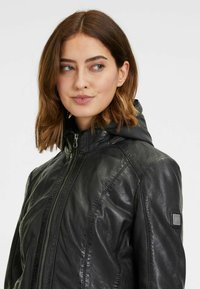 Giacca in pelle nera con chiusura a zip, dotata di cappuccio, cuciture in vista e finitura testurizzata. Include una piccola etichetta con logo sulla spalla.