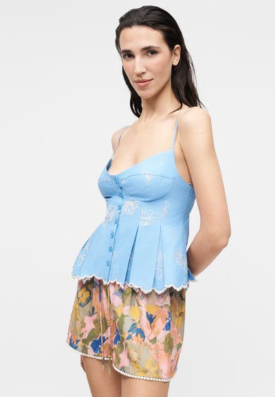 Γυναίκα με σκούρα μαλλιά που φοράει ένα ανοιχτό μπλε αμάνικο τοπ με κεντήματα λουλουδιών και πολύχρωμα λουλουδάτα σορτς με κυματιστές άκρες.