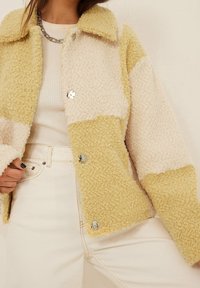 Kurtka polarowa w kolorze kremowym i pastelowym żółtym. Posiada kołnierz, teksturowaną powierzchnię i metalowe guziki. Nosi się ją na jasnej bluzce.