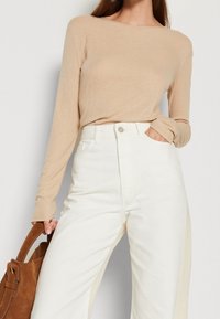Beige geripptes langärmliges Oberteil, kombiniert mit weißen hoch taillierten Hosen. Eine braune Wildledertasche hängt über dem Arm.
