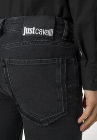 Black denim jeans with frayed edges, a prominent back pocket, and a Just Cavalli logo patch on the waistband. Smooth texture and fitted design.