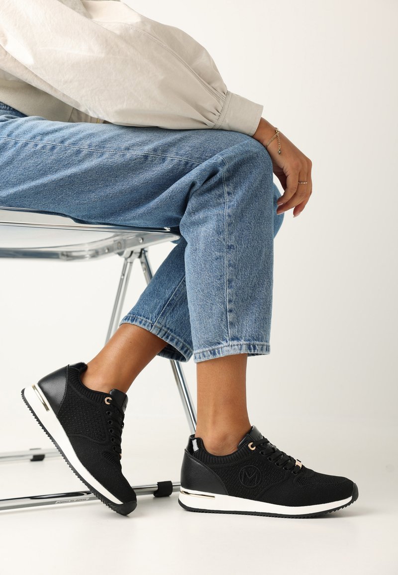 Svart sneaker med en kombination av strukturstickad och lädermaterial, med guldaccenter, rund tå och vit sula.