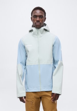 Mężczyzna w jasnoniebieskiej i blado szarej bluzie z kapturem i zamkiem błyskawicznym oraz brązowych spodniach, stoi na jednolitym białym tle.