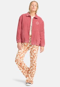 Giacca di velluto rosso con collo e polsini elasticizzati, abbinata a pantaloni crema con un motivo floreale astratto marrone.
