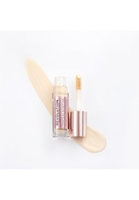 Correcteur Revolution Conceal & Define dans un tube transparent, avec un applicateur doux, présente une formule crémeuse, beige clair, testée à côté.