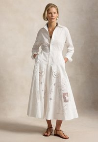 Femeie purtând o rochie albă lungă cu nasturi, decorată cu modele sculptate complexe, stând cu mâinile în buzunare, asortată cu sandale maro.