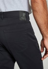 Zwarte broek van een gestructureerde stof, met zichtbare stiksels, een achterzak en een label met "JP 1880" in een rechthoekig ontwerp.