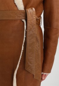 Abrigo de cuero marrón con forro de borrego, detalles de costuras verticales y un cinturón atado en la cintura. Textura suave con acabado mate.