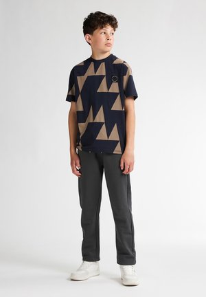 T-shirt marine avec un motif géométrique beige, manches courtes, col rond ; associé avec un pantalon de jogging gris et des baskets blanches.