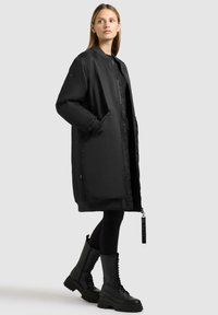 Cappotto imbottito nero con design bomber, maniche lunghe, tasche laterali e colletto piatto. Abbinato a stivali militari neri con lacci.