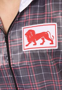 Karirana jakna na zip ima crno-bijelo-crvenu šemu; uključuje istaknutu crvenu lionsku zakrpu na bijeloj podlozi.