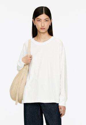 White long-sleeve t-shirt in soft fabric with a relaxed fit, paired with a beige suede shoulder bag. Dark denim jeans are also worn.