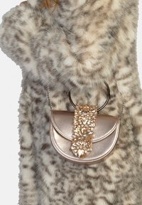 Borsa a mano in oro metallizzato dalla forma curva, decorata con una fascia di pietre preziose trasparenti e una tracolla a catena, su uno sfondo di pelliccia testurizzata.