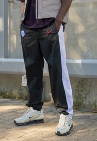 Sorte og hvide joggers har en sidelås, Nike-logo og et Paris Saint-Germain-patch, parret med hvide og sorte sneakers.