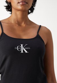 Black tank top made of soft cotton, featuring thin straps and a white "Calvin Klein Jeans" logo across the front. Smooth texture.