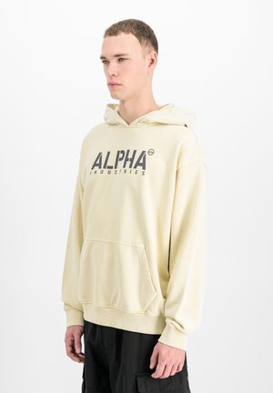 Giovane uomo con felpa con cappuccio color crema Alpha Industries e pantaloni cargo neri, in piedi davanti a uno sfondo bianco semplice, rivolto a sinistra.