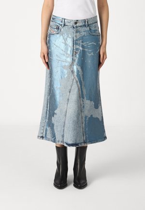 Femme portant une longue jupe en denim bleu métallique lavé à l'acide et des bottines en cuir noir, debout devant un fond uni.