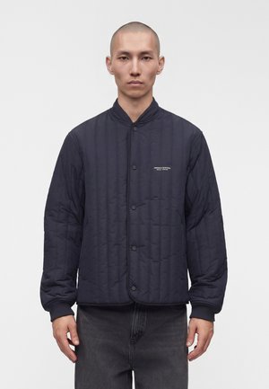 Ung mand med barberet hoved iført en sort quiltet bomberjakke med knapper foran og Armani Exchange-logo på brystet, stående mod hvid baggrund.