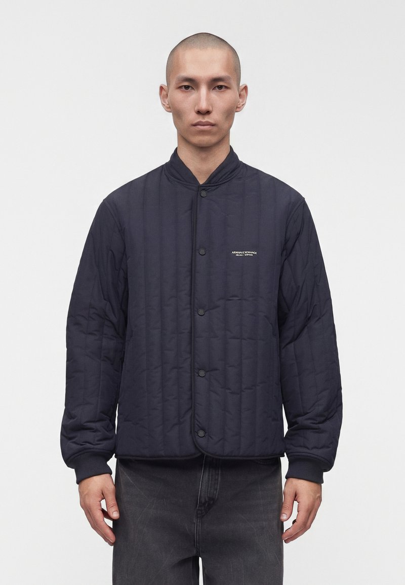 Jeune homme au crâne rasé portant une veste bomber matelassée noire avec des boutons à l'avant et le logo Armani Exchange sur la poitrine, debout sur un fond blanc.